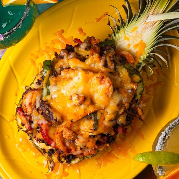 Pineapple stuffed with melted cheese, shrimp, and vegetables on a yellow plate with a margarita.