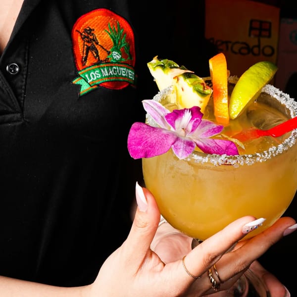 A server in a Los Magueyes uniform holds a large, garnished margarita with pineapple, lime, and an orchid.