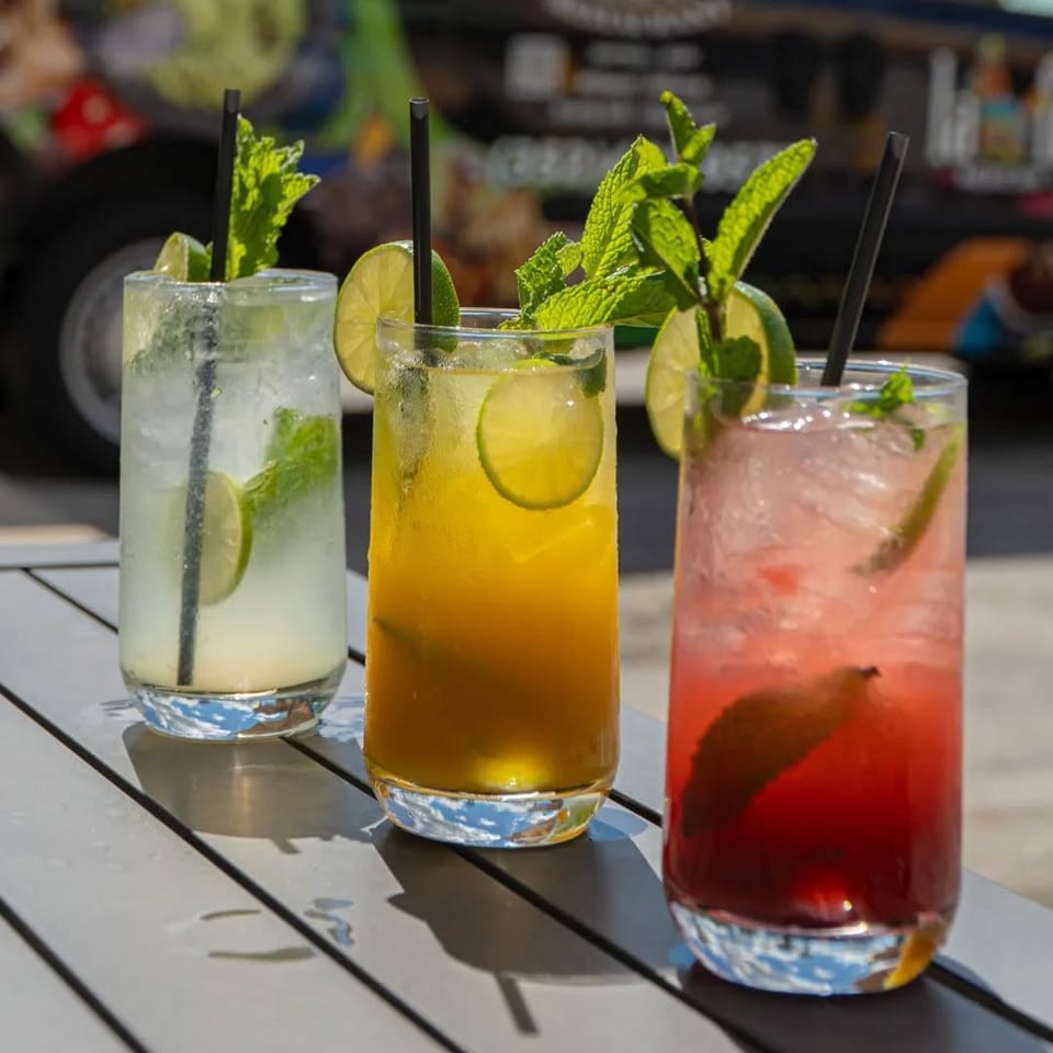 Three colorful Mexican cocktails, garnished with lime and mint, sit on a gray outdoor table.