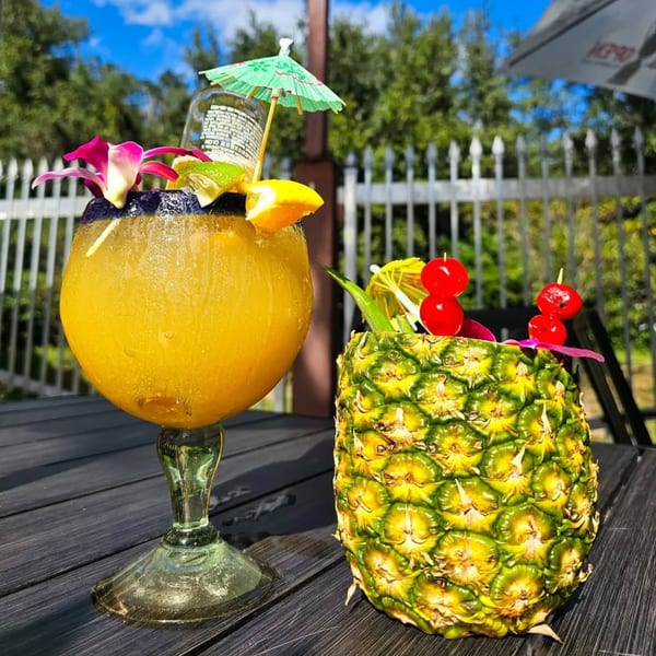 A large yellow margarita with a beer bottle and a pineapple drink with cherries on a dark table.