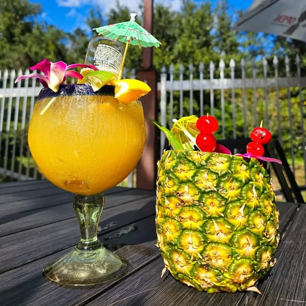 A large margarita with a beer bottle and a pineapple drink with cherries on a dark outdoor table.