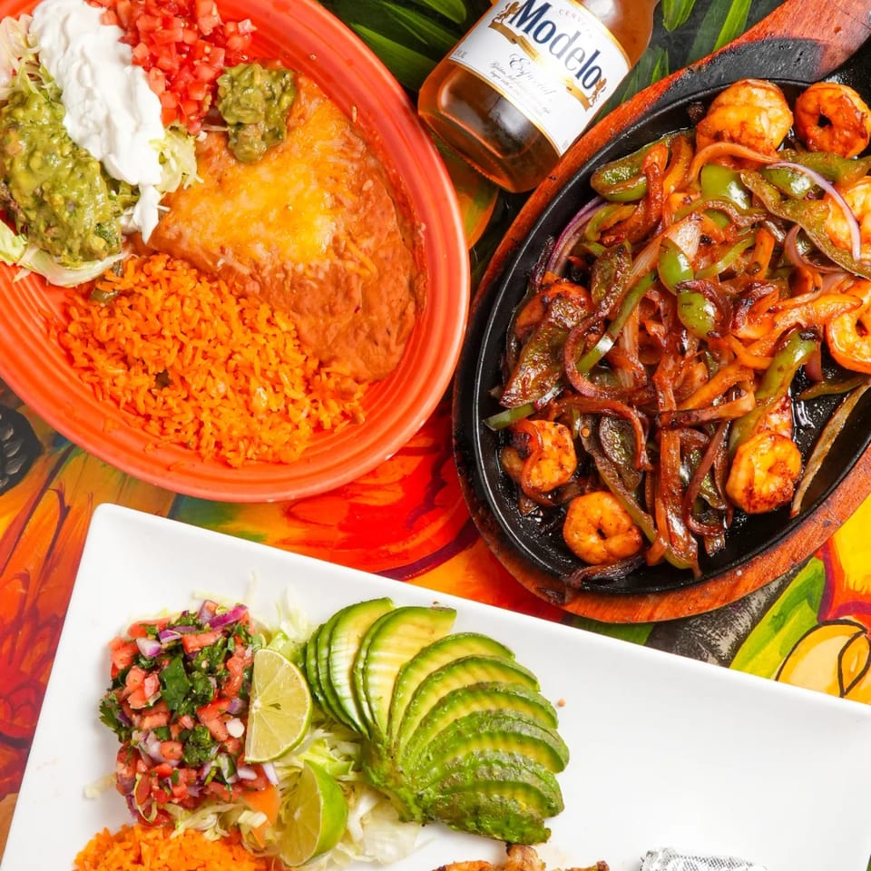 A vibrant Mexican feast featuring shrimp fajitas, a cheesy enchilada, rice, guacamole, and a Modelo beer.