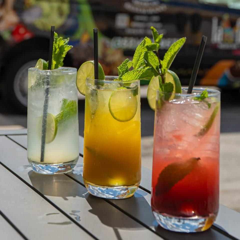 Three colorful mojitos, garnished with lime and mint, sit on a table outdoors.