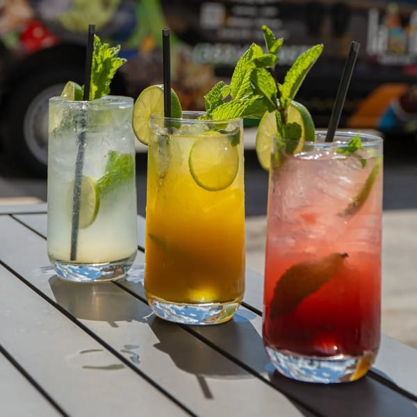 Three colorful mojitos, garnished with lime and mint, sit on a gray outdoor table.