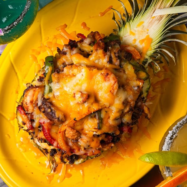 Grilled pineapple half filled with melted cheese, shrimp, and vegetables on a yellow plate.