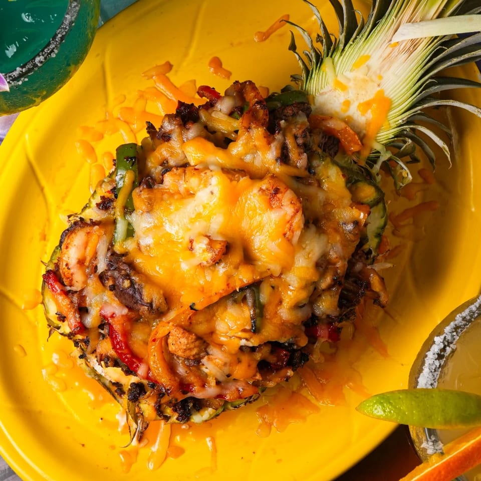 Grilled pineapple half filled with melted cheese, shrimp, and vegetables on a yellow plate.