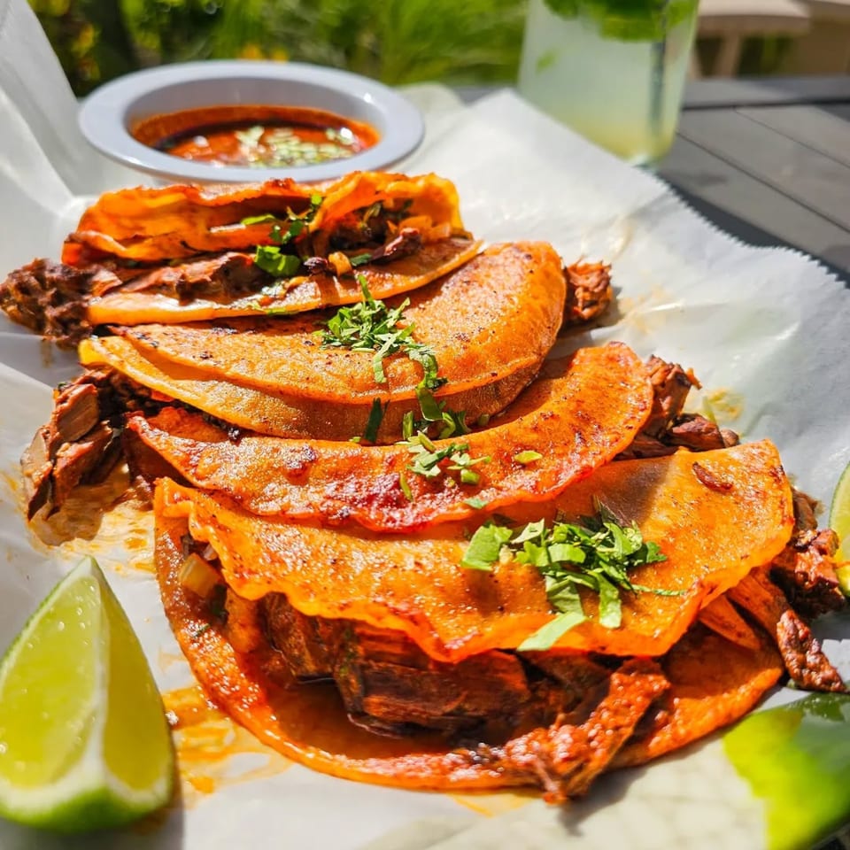 Los Magueyes birria tacos with cilantro, lime wedges, and a side of consommé.