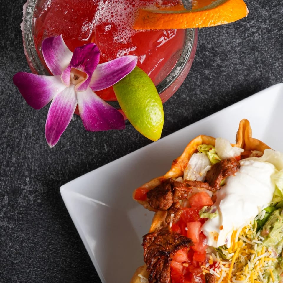 A vibrant red margarita with an orchid and lime, next to a taco salad with meat, lettuce, and sour cream.